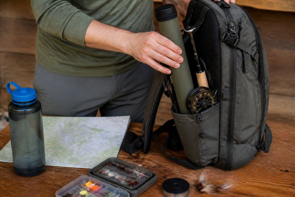 Hands organizing fishing tackle, rod tube, and reel into a backpack's side compartments, with a topographic map and water bottle visible nearby
