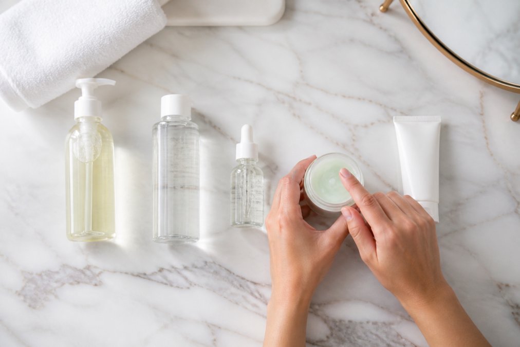 Skincare products arranged in application order on a white marble surface from left to right (cleanser, toner, serum, lightweight moisturizer, sunscreen), with hands demonstrating the layering sequence by holding a gel moisturizer