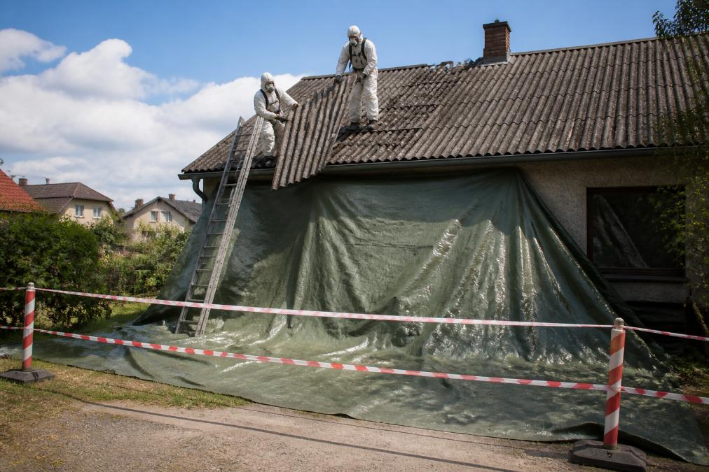 Pracovníci v bílých ochranných oblecích a respirátorech demontují eternitové šablony ze střechy rodinného domu, pod nimi zakryté plachty a vyznačená bezpečnostní zóna, denní světlo