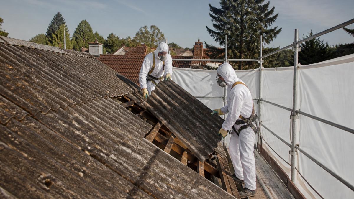 Pracovníci v bílých ochranných kombinézách, respirátorech a rukavicích demontují eternitové šablony ze střechy rodinného domu, kolem je postavené lešení a ochranná fólie, denní světlo