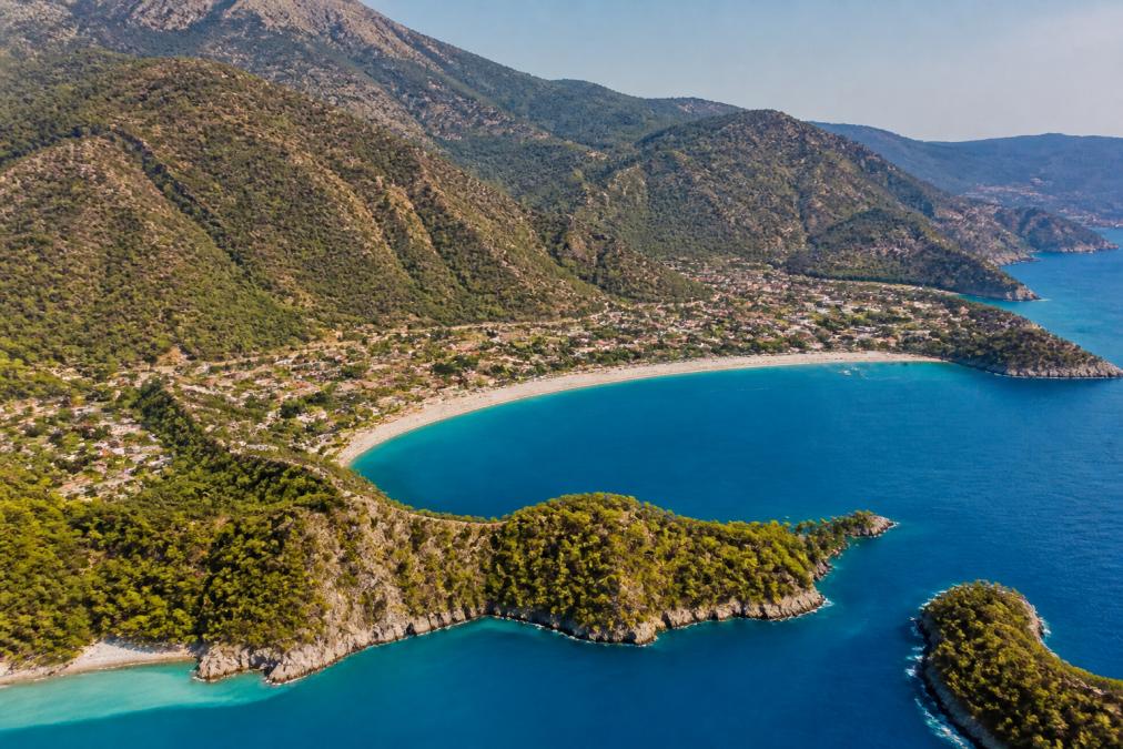 Aerial view of Ölüdeniz bay with turquoise waters, green mountains in background, and coastal settlements nestled below, showcasing the natural beauty of the Lycian Way starting point