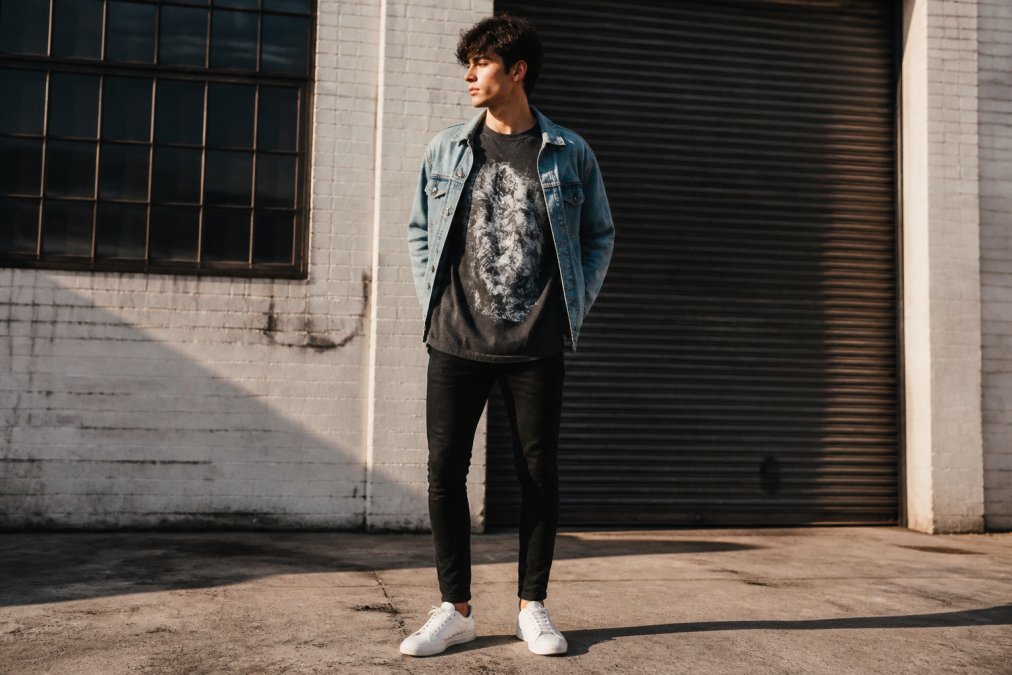 Person wearing an oversized heavyweight graphic tee layered with an open denim jacket, styled with fitted black jeans and white minimal sneakers, standing confidently in urban setting with natural afternoon sunlight