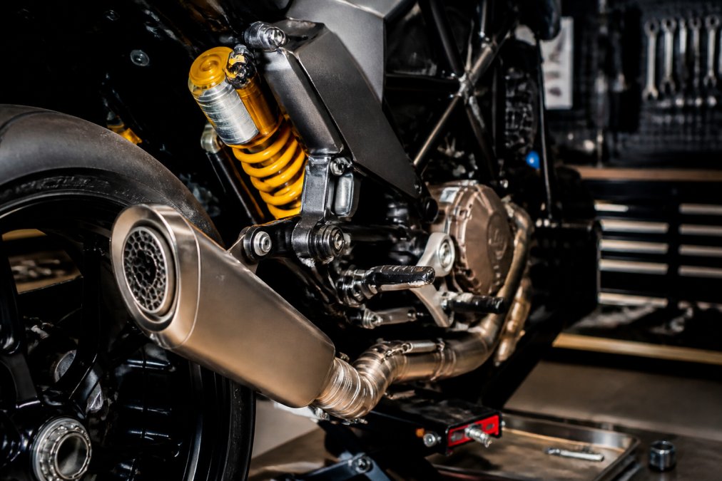 Close-up van een Husqvarna motorfiets met zichtbare Ohlins-vering en SC-project uitlast in een werkplaats met professionele verlichting, technische details onder heldere werklampen