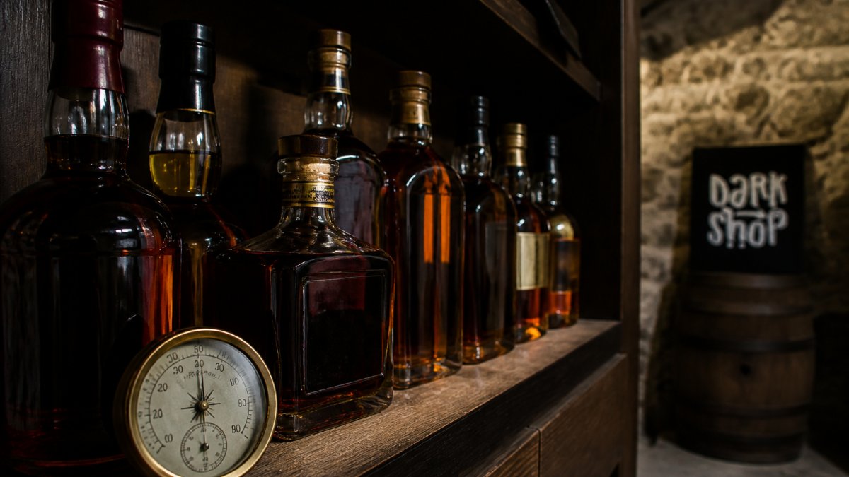 Butelki whisky stojące pionowo na ciemnej drewnianej półce w chłodnej piwnicy, z dala od światła słonecznego, obok higrometru