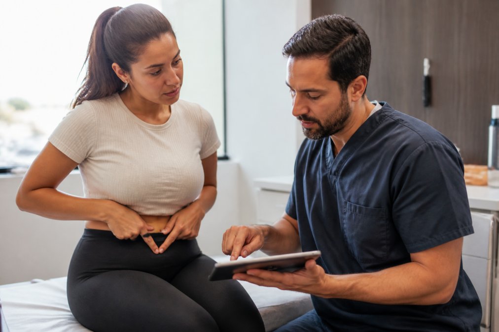 A patient in a consultation room with a board-certified plastic surgeon, pointing to their midsection while reviewing a treatment plan on a tablet, soft natural lighting, professional medical setting