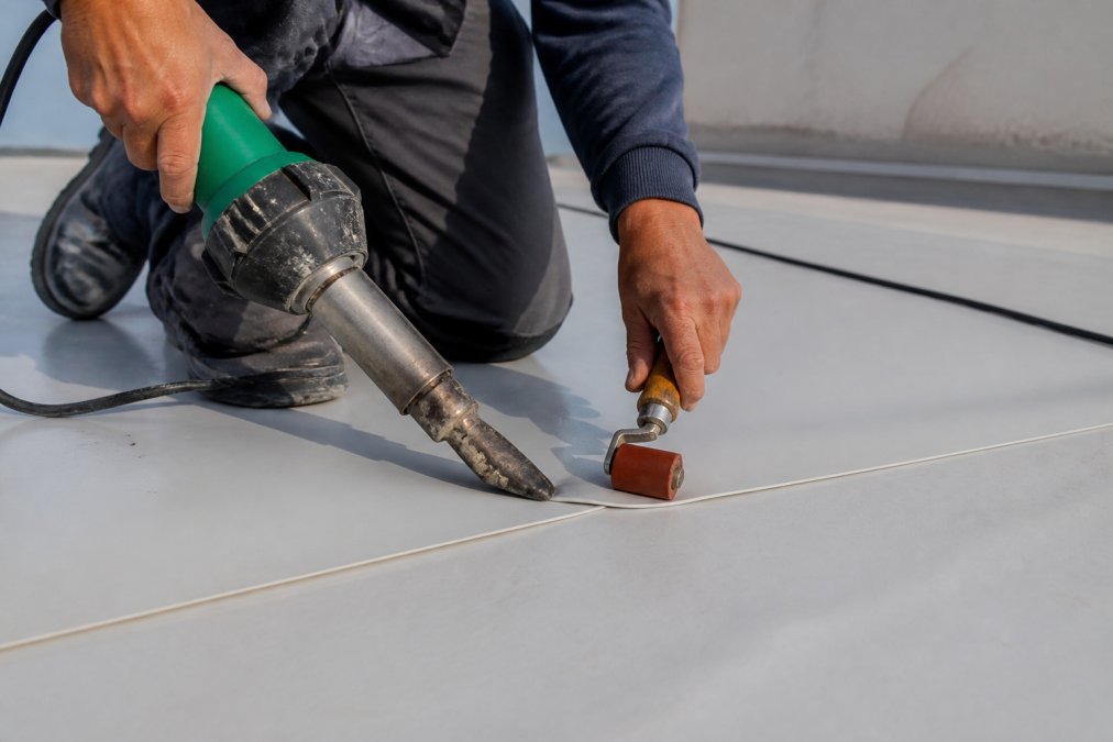 Close-up of technician's hands performing heat-welding on a single ply TPO membrane seam using specialized heat gun equipment, with seam being fused together under bright workshop lighting