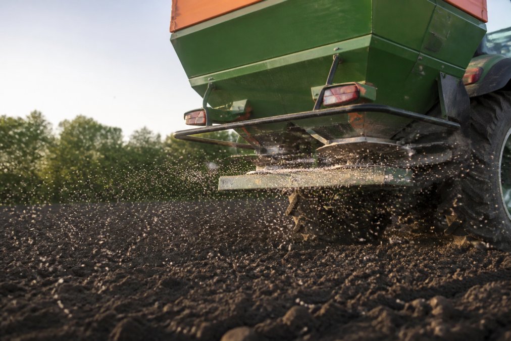 Close-up of granular fertiliser pellets being spread across dark arable soil from a broadcast spreader attachment on a tractor, in soft morning light