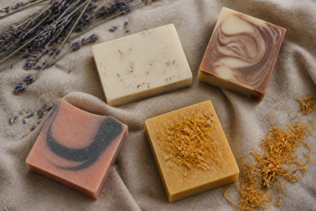 Overhead flat lay of four artisan soap bars with different colors and textures arranged on natural linen cloth with dried lavender and calendula scattered nearby, warm natural lighting from above