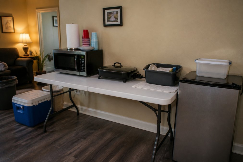 A neatly organized temporary kitchen setup on a folding table in a living room, featuring a microwave, electric skillet, mini-fridge, paper plates, and a small dish tub for washing, under warm indoor lighting