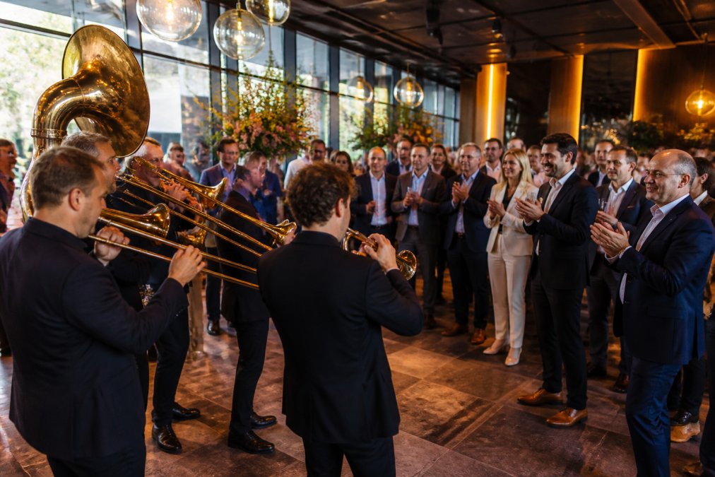 Een professionele brassband in donkere pakken speelt tijdens een chique bedrijfsreceptie in een moderne evenementenlocatie, gasten in zakelijke kleding kijken toe en klappen mee, warme sfeerverlichting