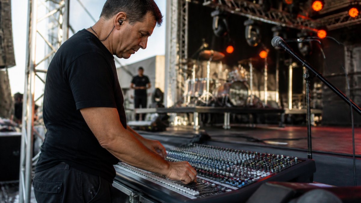 Professional audio technician setting up mixing console and testing microphone levels at live event venue with stage lighting rigs visible in background