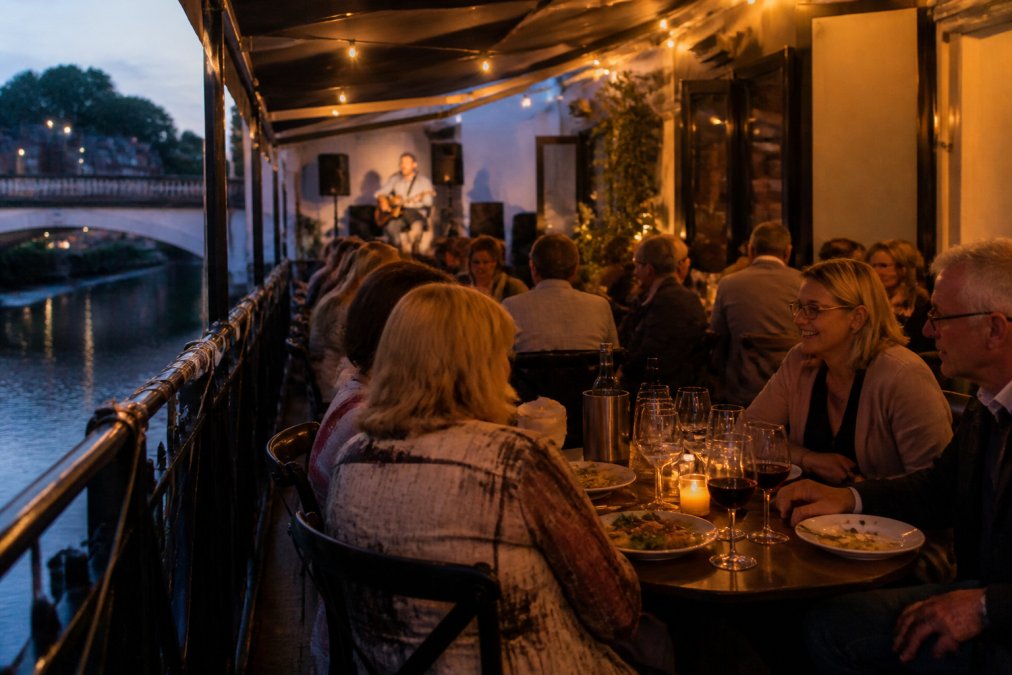 Candlelit tables set along the riverside with diners enjoying meals, warm ambient lighting reflecting off wine glasses, live performer visible on a small stage in background