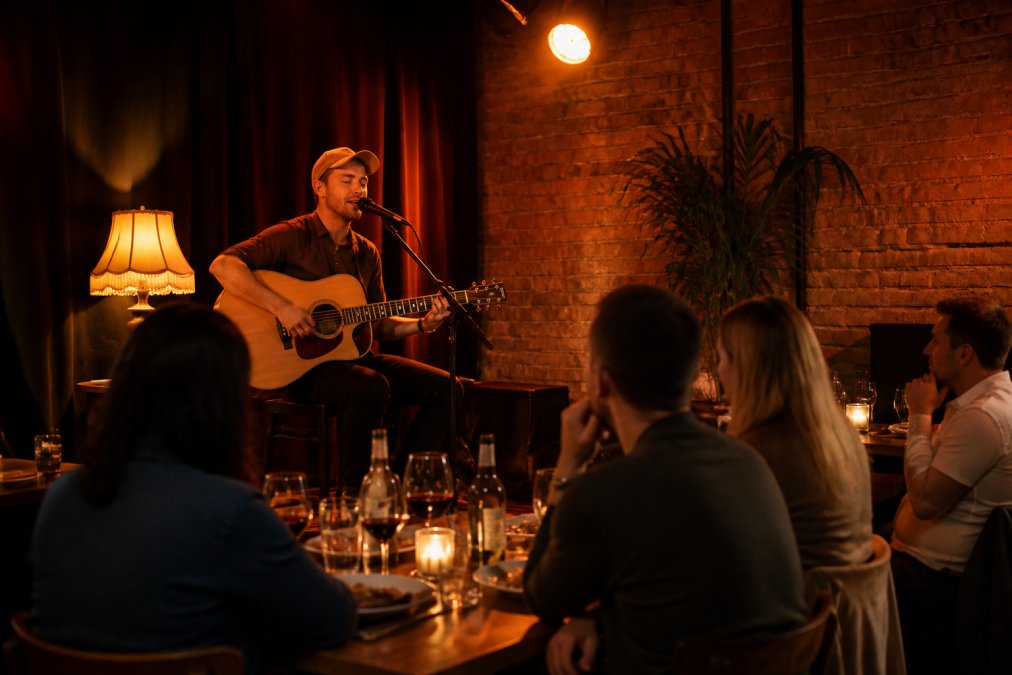 Intimate live performance scene with acoustic musician playing guitar on small stage, warm amber stage lighting, diners seated at nearby tables with wine glasses, cosy restaurant ambiance