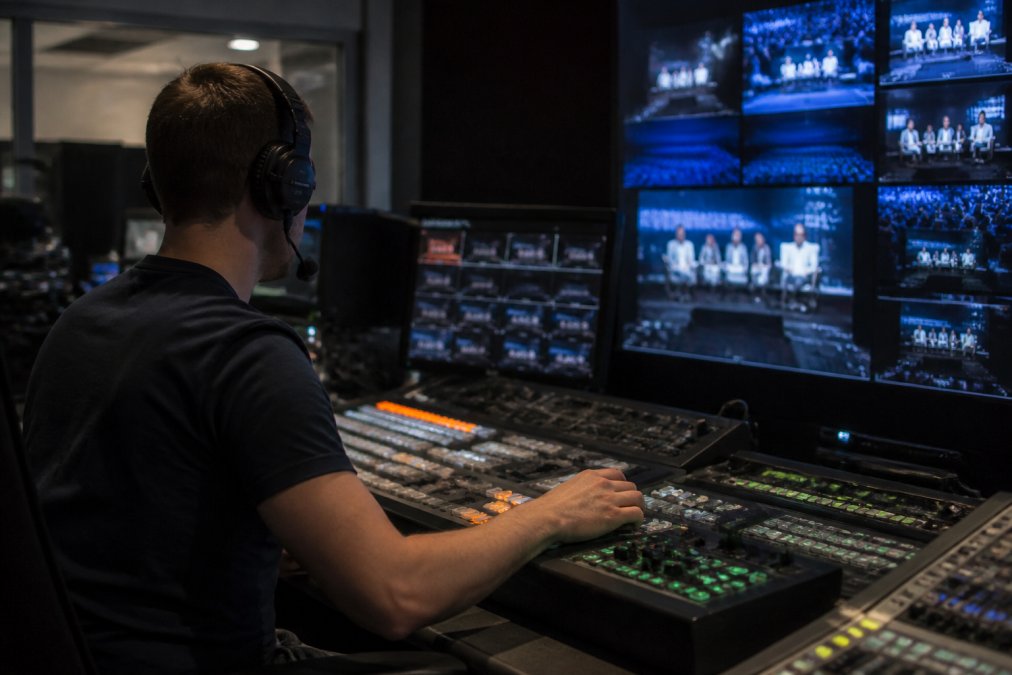 A technician in a broadcast control room monitoring multiple screens showing live video feeds, dim lighting with the glow of monitors reflecting on the console