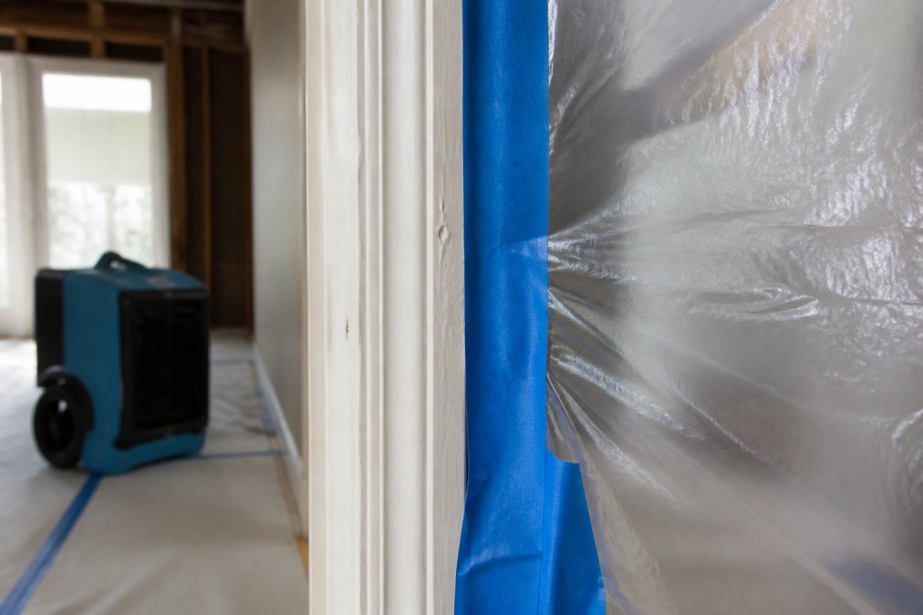 Close-up of blue painter's tape sealing plastic sheeting along doorframe edge, with transparent plastic barrier creating visible separation between construction zone and living space, HEPA air filtration unit visible in soft-focused background