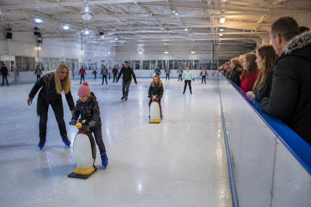 Families enjoying an indoor ice skating session, children holding penguin skate aids on the ice, bright rink lighting, spectators watching from the side