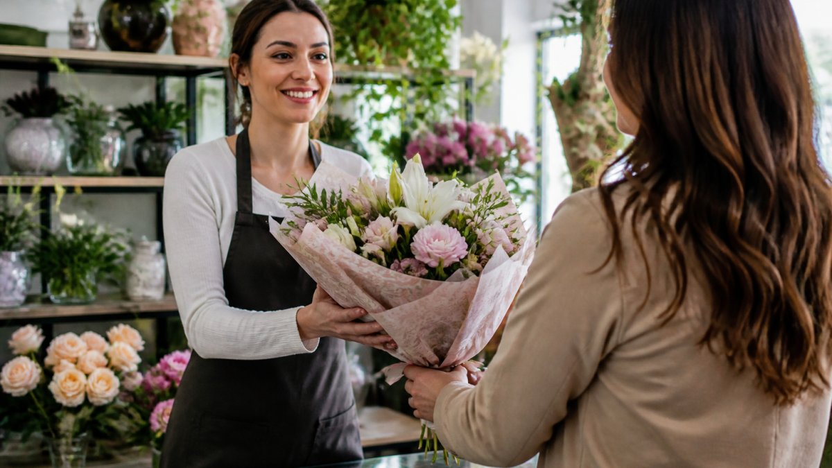 A smiling florist in an apron handing a wrapped bouquet of fresh flowers to a customer inside a bright, well-stocked local flower shop