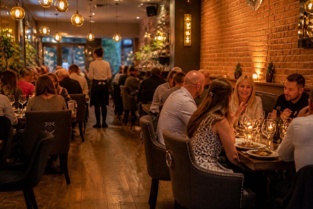 Vibrant restaurant interior with diners enjoying meals at candlelit tables, warm golden lighting creating a welcoming atmosphere with exposed brick walls and modern décor