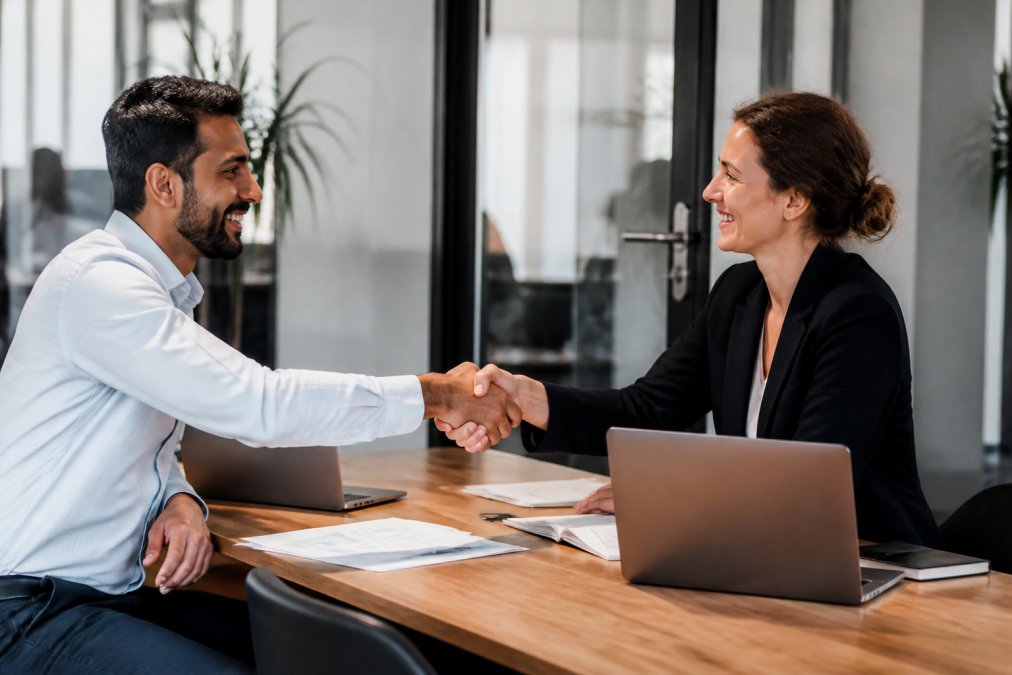 Two business professionals from different cultural backgrounds shaking hands across a meeting table, with laptops and documents visible, in a modern European office setting