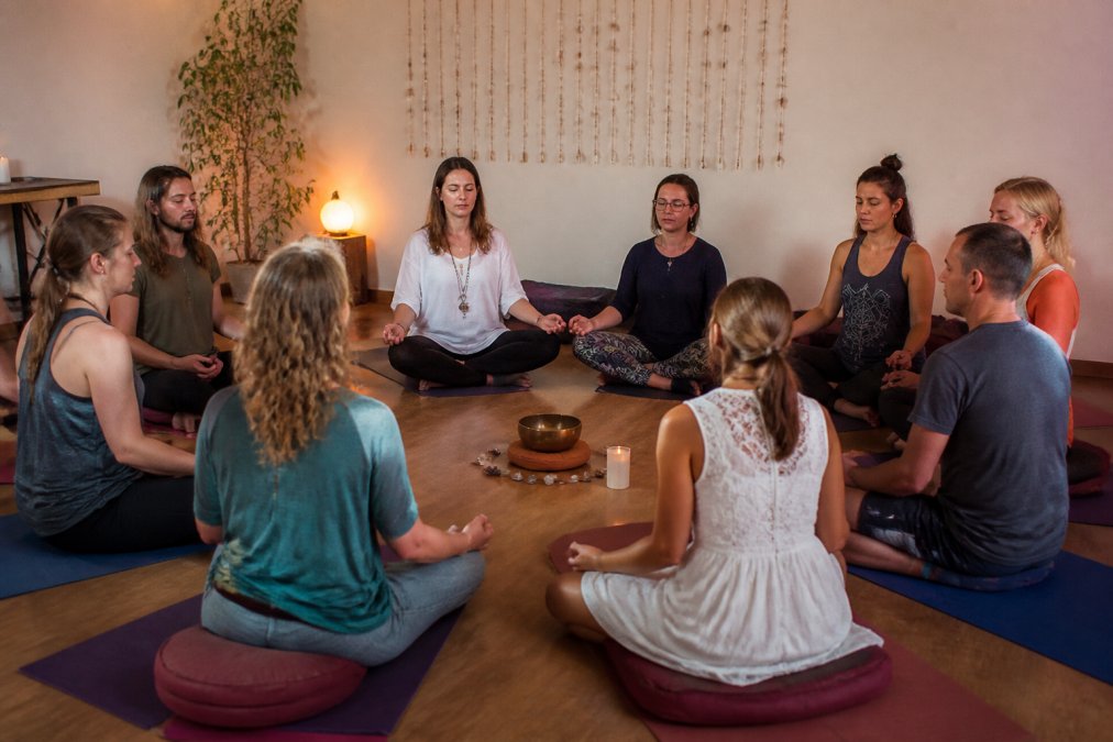 A diverse group of adults sitting in a circle on yoga mats and cushions in a softly lit room, eyes closed in meditation, with a singing bowl in the center