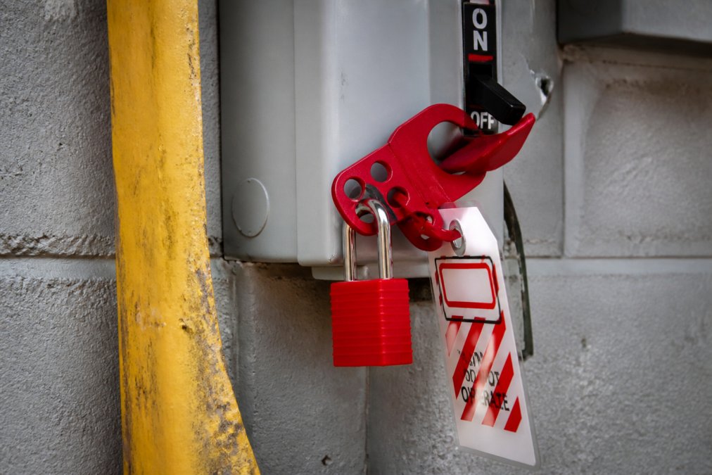 Close-up of a red safety padlock and lockout hasp attached to an industrial electrical disconnect switch on a factory wall, with a warning tag visible