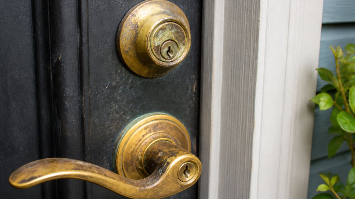 Close-up of a residential deadbolt and door handle showing visible wear, corrosion, and tarnishing on the lock cylinder and surrounding brass hardware under natural daylight