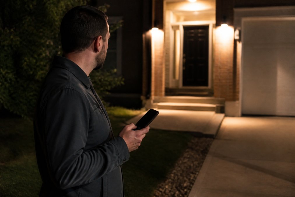 Homeowner standing in a well-lit area outside their house at night, calmly checking their phone for emergency contact information while looking toward their front door in the distance