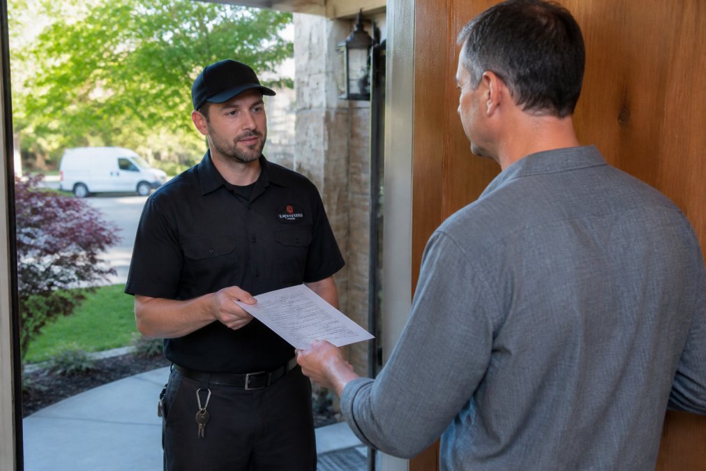 A professional locksmith in a dark uniform handing a clear, itemized paper invoice to a homeowner at a front door, natural daylight, suburban setting