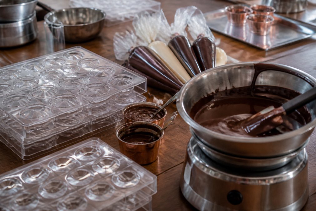 Close-up of professional chocolate-making equipment neatly arranged on a workshop table: polycarbonate moulding trays, piping bags, small pots of ganache, and a tempering bowl with glossy dark chocolate, ready for a hands-on session