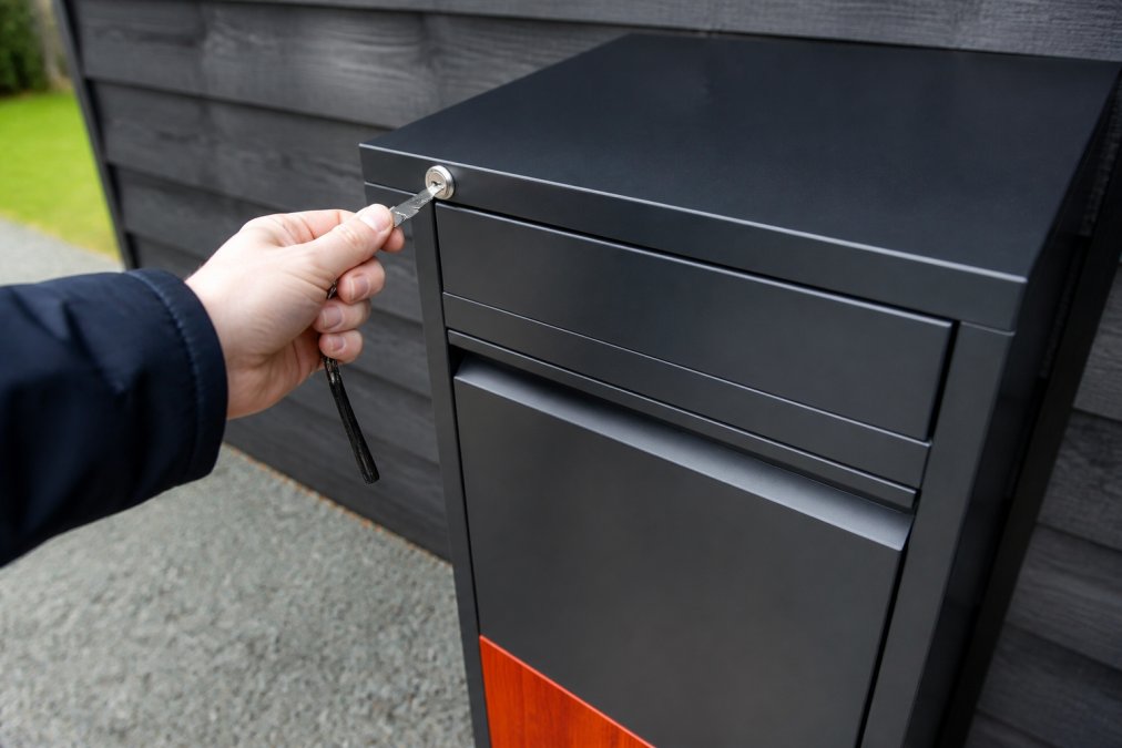 Close-up van cilinderslot en beveiligingsmechanisme op pakketbrievenbus, hand die sleutel toont, details van roestvrij staal zichtbaar