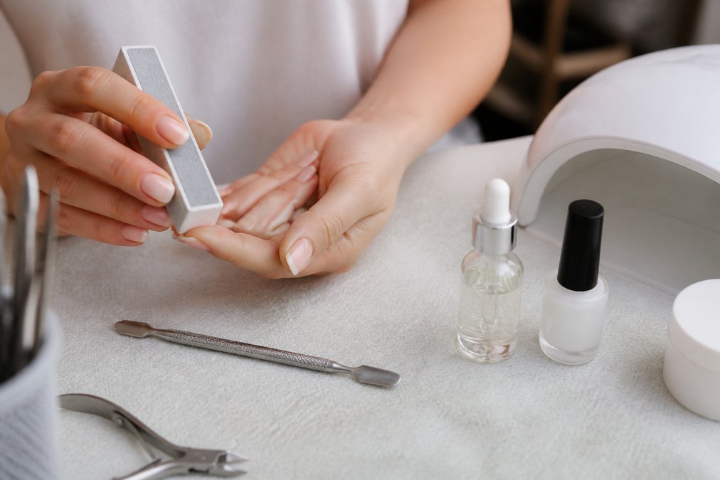 Close-up of a woman's hands using a nail buffer and cuticle pusher on a clean manicure station with bottles of dehydrator and primer visible