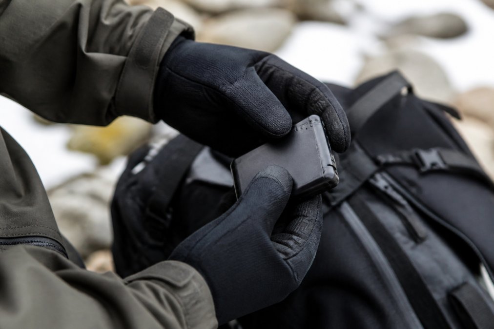 Close-up of a person's hands wearing low-profile heated gloves, holding a lithium-ion battery pack and examining the mAh rating label, with outdoor winter clothing visible in the background