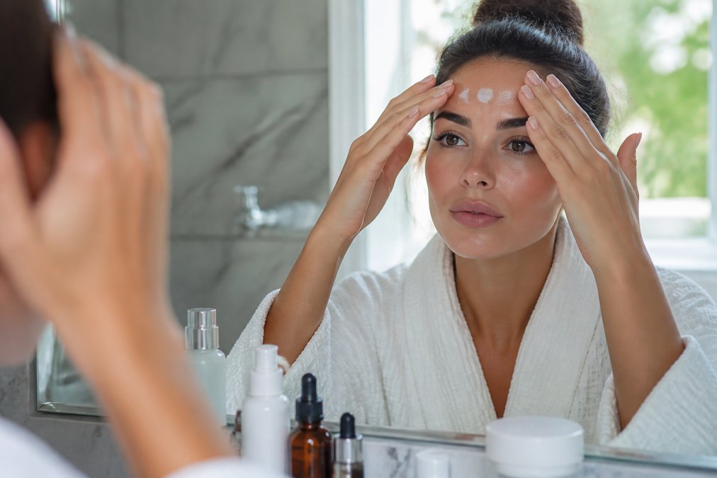 Close-up of hands applying sunscreen to the forehead and brow area, with skincare products visible on a marble bathroom counter, morning natural light through a window