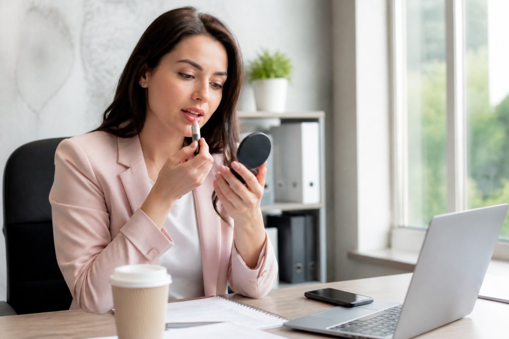 A professional woman in a bright office applying a nude pink lipstick using a small mirror at her desk, with a laptop and coffee cup visible nearby