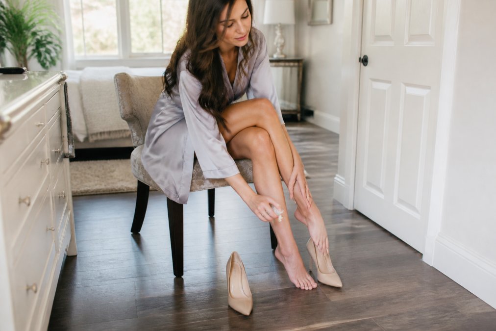 A woman in a bright modern bedroom sitting on the edge of a chair, spraying a cooling mist onto her foot before slipping on elegant nude high heels, soft natural morning light through the window