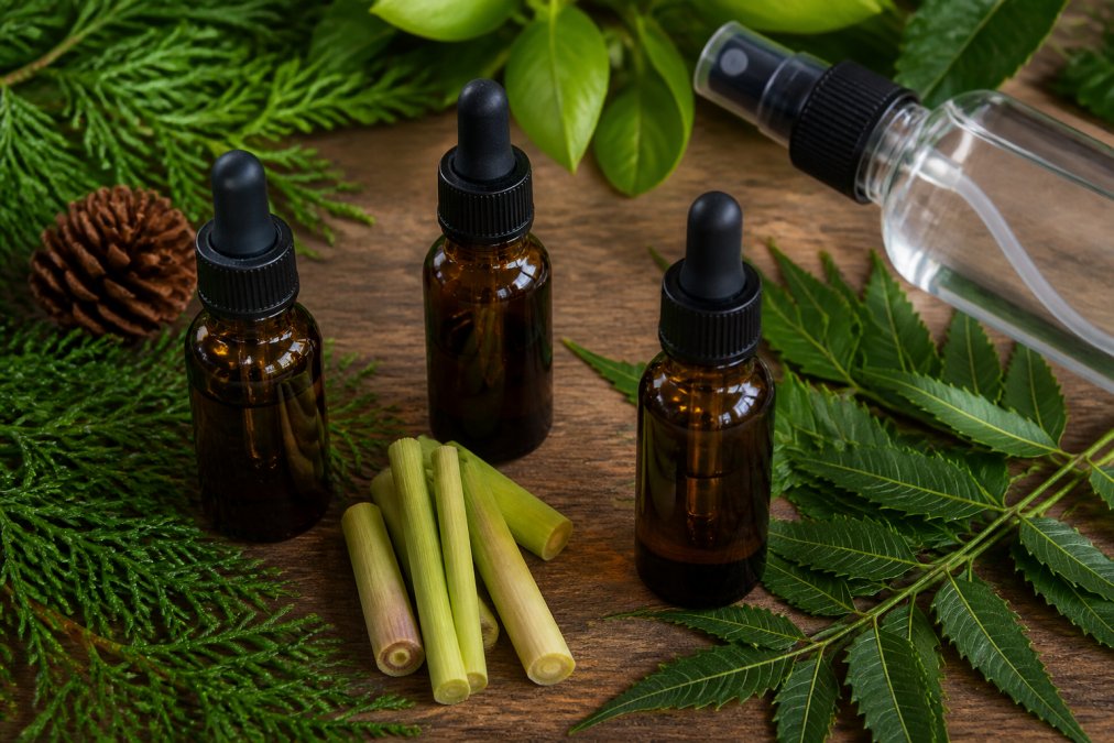 Close-up flat lay of small amber dropper bottles filled with essential oils, cedar, lemongrass, and neem, arranged on a wooden surface with green leaves and a spray bottle nearby