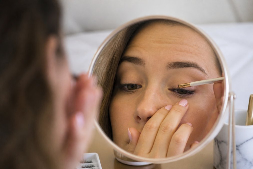 Close-up of hands carefully applying pre-glued lash cluster under the natural lash line using tweezers, showing proper under-the-lash placement technique with magnified mirror reflection visible