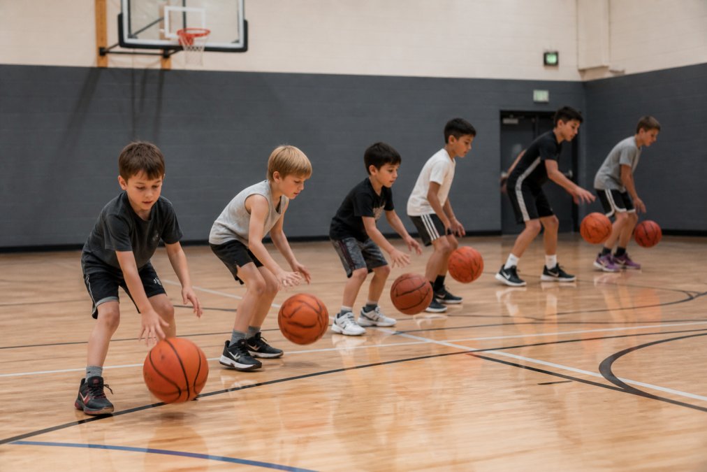 Young basketball players of mixed ages practicing ball-handling drills on a court, showing progression from younger children dribbling at slower pace to older kids executing more complex moves
