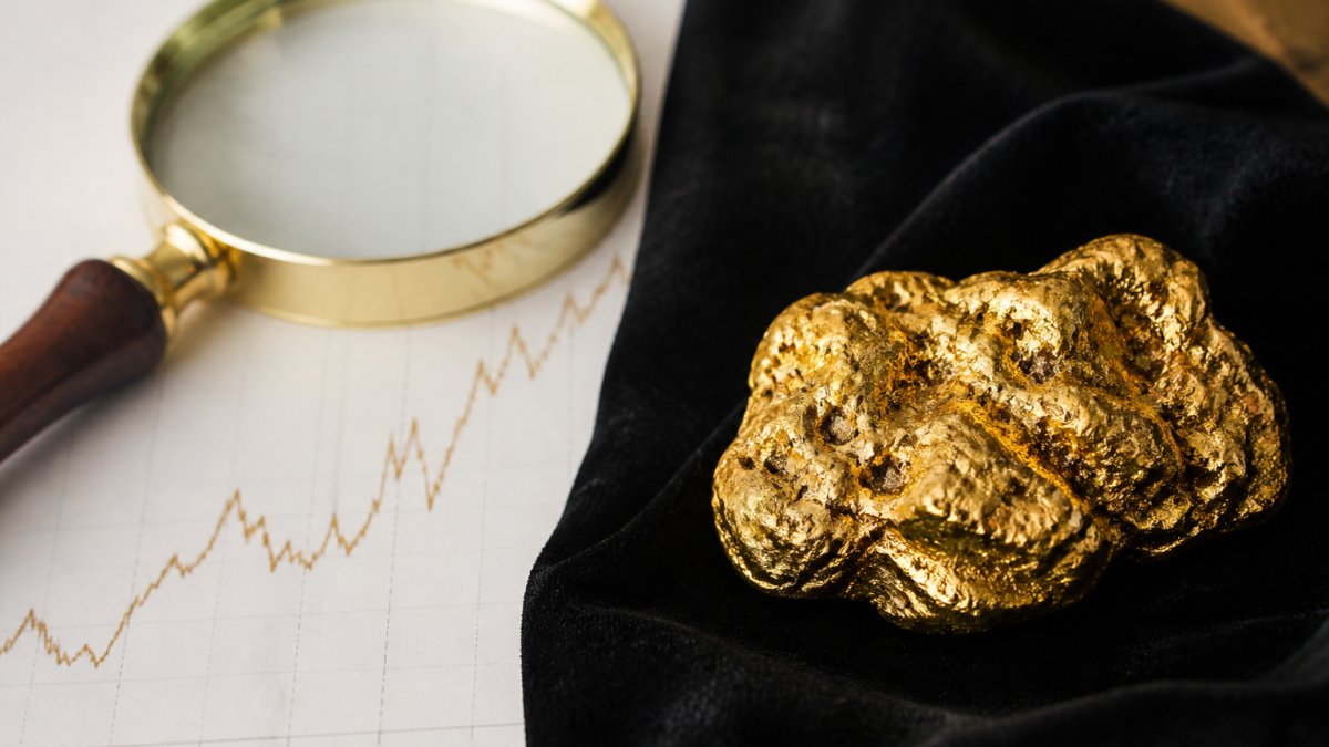 A close-up of a natural Australian gold nugget resting on dark velvet beside a printed price chart and a magnifying glass, warm side lighting