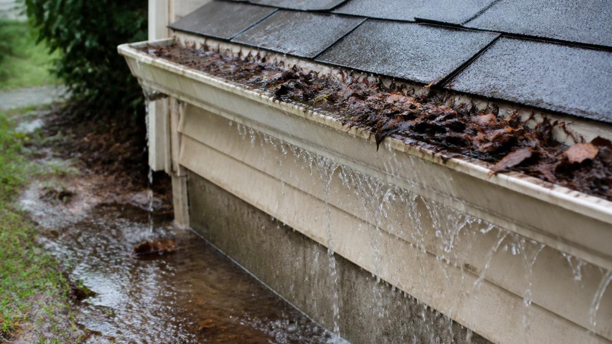 Close-up of a clogged gutter filled with leaves and debris, water overflowing down the side of a house near the foundation during heavy rain