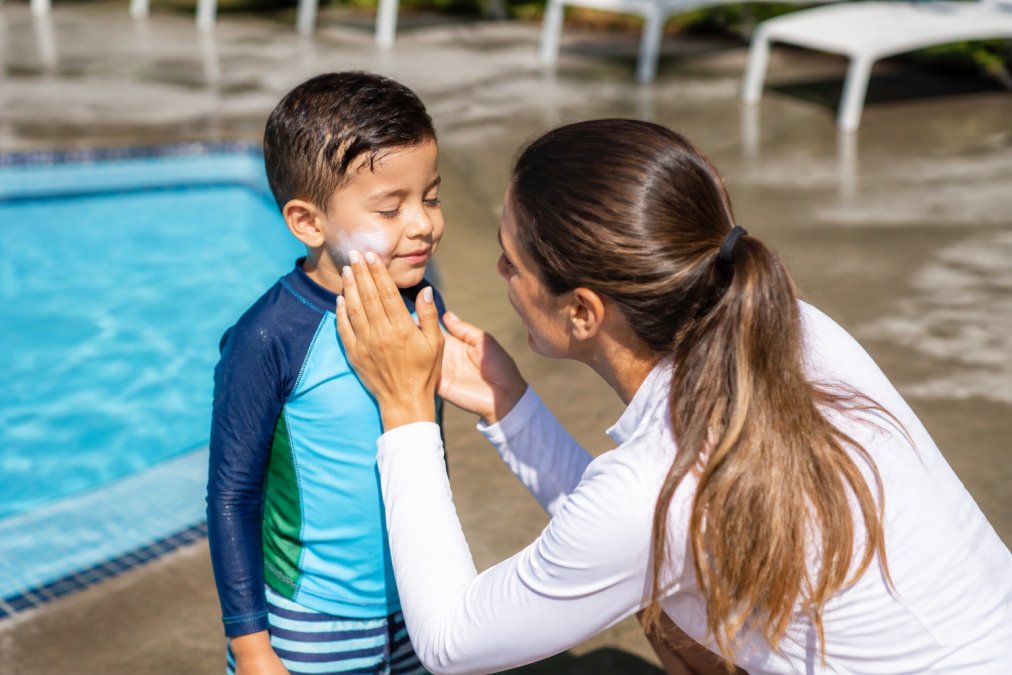 A parent applying sunscreen to a young child's face while the child wears a long-sleeve rash guard swimsuit at a sunny outdoor pool, bright midday light, wet pool deck in the background