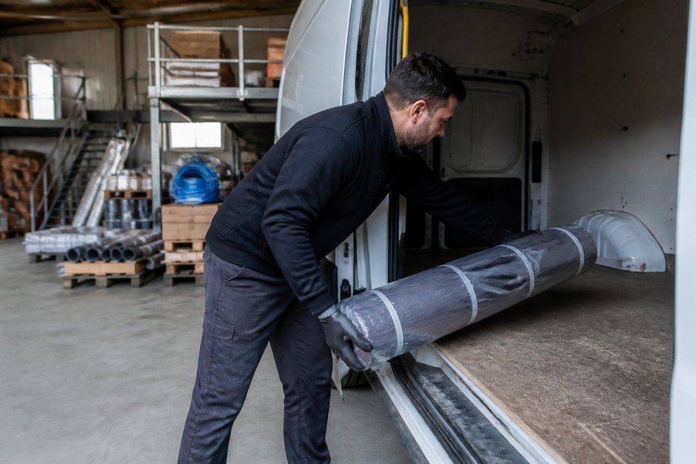 Pakketje met lood op rol wordt voorzichtig in een bestelwagen geladen op een professionele werkplek met opslag en gereedschap zichtbaar