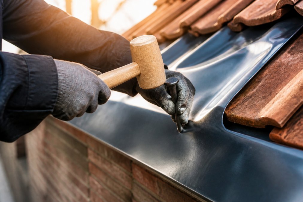 Professionele dakdekker die bladlood voorzichtig tegen dakrand bevestigt met houten hamer en loden klopper, zonlicht schijnt op het werkstuk
