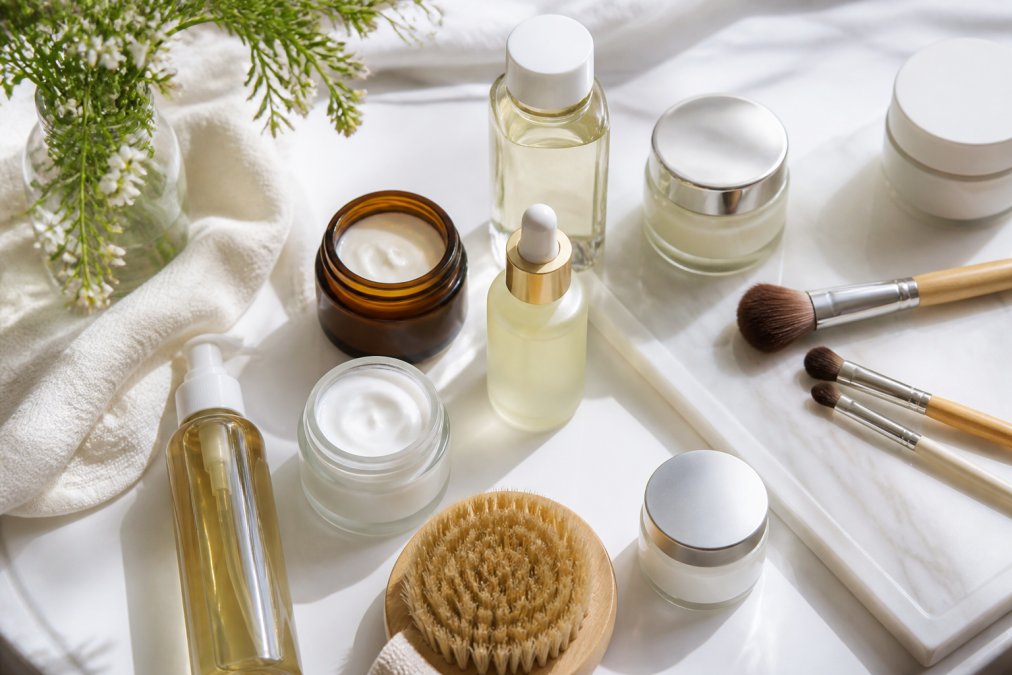 A flat lay of a carefully arranged selection of premium skincare products and makeup brushes on a clean, minimalist white surface, soft natural light casting gentle shadows