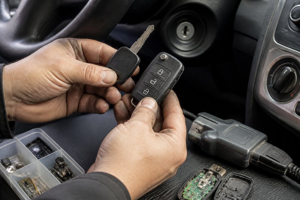 Close-up of hands holding a modern transponder car key and smart key fob next to a vehicle's ignition, with the key blank, microchip components, and diagnostic scanner visible