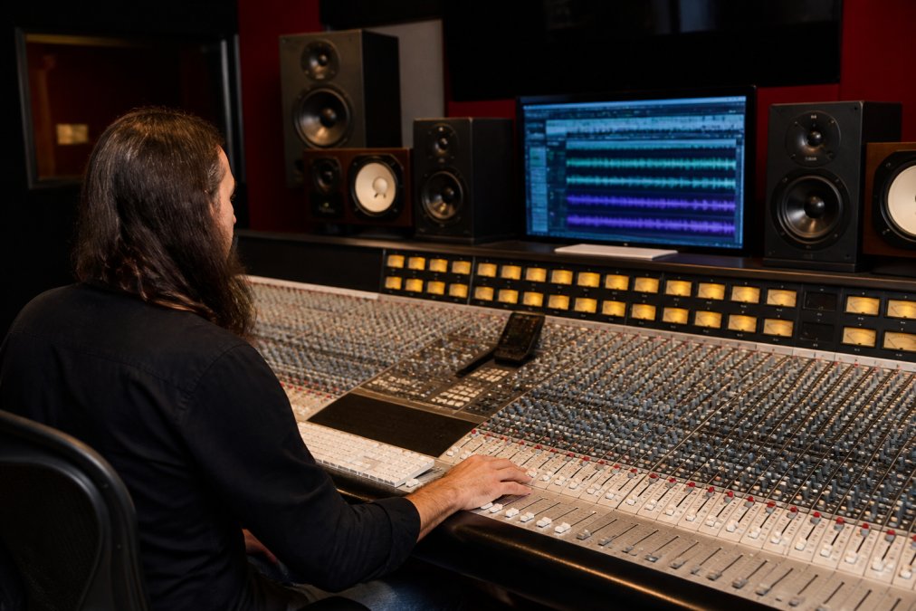 A mastering engineer seated at a large analog console in a dimly lit studio, hands on faders, with large studio monitors in the background and a DAW screen showing waveform displays