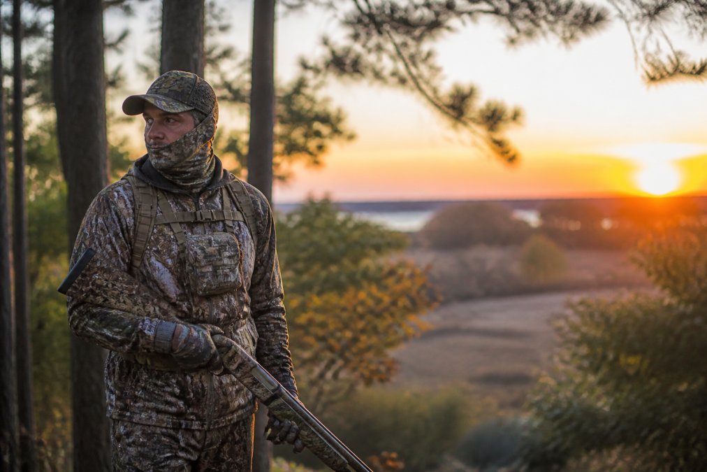 Hunter in full camouflage gear standing in a wooded area at dawn, wearing quality hunting apparel and demonstrating proper field preparation and readiness