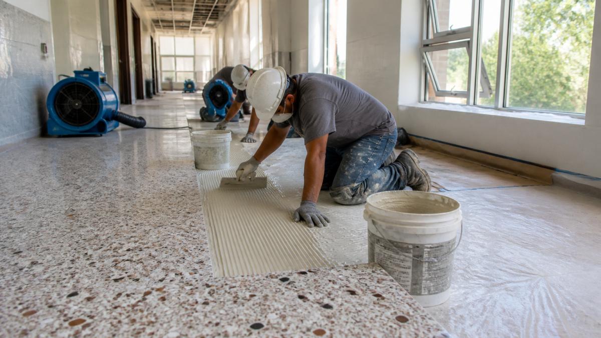 Professional flooring installation team applying low-VOC adhesive to new terrazzo flooring in a campus hallway, with industrial ventilation equipment running in the background and windows open for air circulation