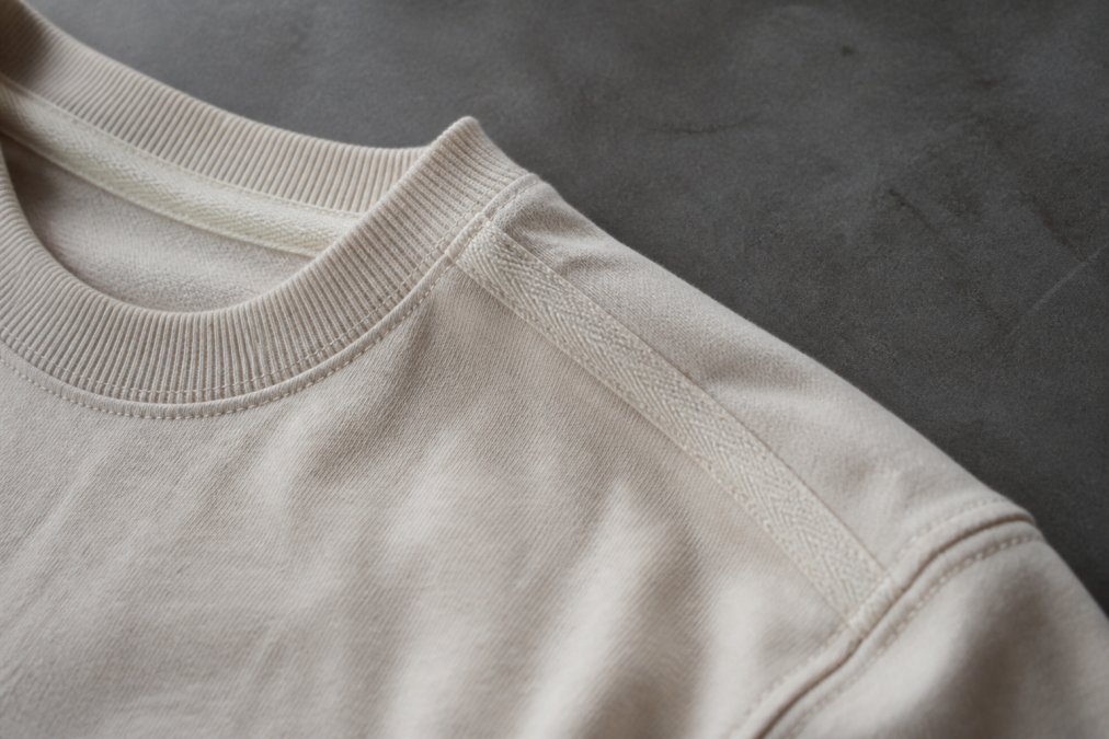 Close-up of high-quality cotton t-shirt fabric showing twill tape reinforcement on shoulders, ribbed collar construction, and stitching detail under natural studio lighting