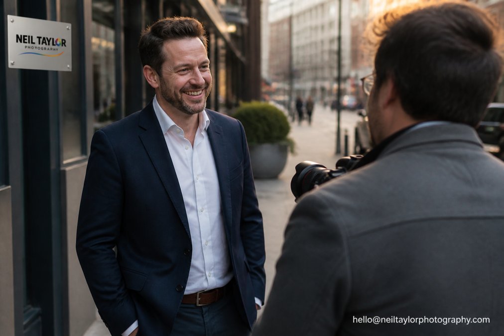 Business professional photographed in natural daylight on urban location with soft-focus professional environment visible in background, confident posture and authentic expression